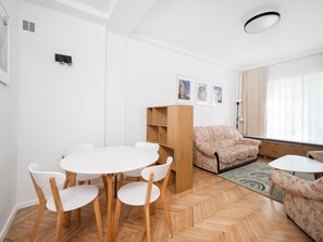 A modern living room with a round table and chairs, a sofa, a wooden bookshelf, and a large window with curtains. - Manufaktura Cosy Apartment (Łódź)