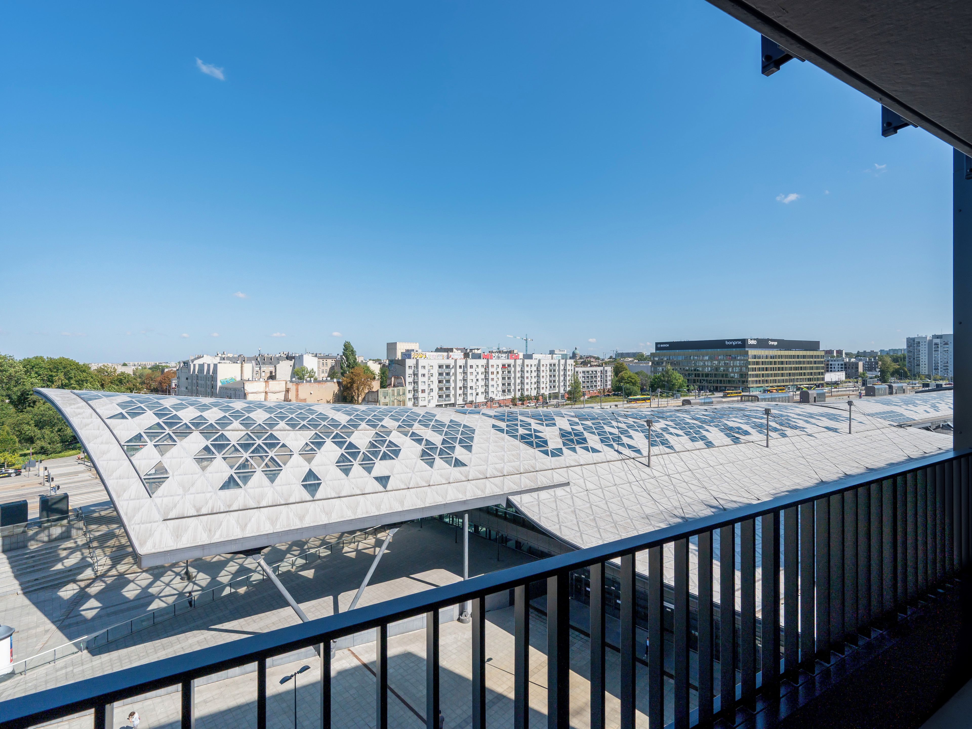 A modern building with a unique geometric roof design, a clear sky, and a view of surrounding urban buildings.