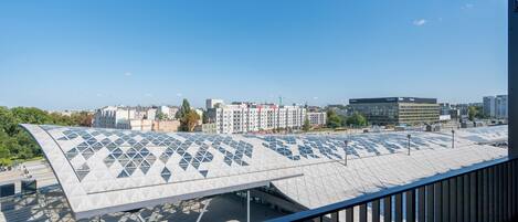 Um edifício moderno com um design geométrico distinto no telhado, céu claro e vista de edifícios urbanos ao redor.