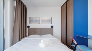 A neatly made bed with white linens and a rolled towel, a headboard, two wall-mounted clocks, and a blue accent wall.