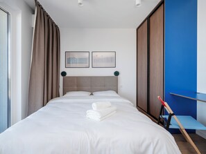 A neatly made bed with white linens and a rolled towel, a headboard, two wall-mounted clocks, and a blue accent wall.