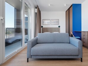 A modern bedroom with a grey sofa, a bed with a wooden headboard, a large window with curtains, and a blue accent wall. - Łódź Fabryczna Modern Studio (Łódź)