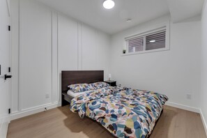 A bedroom with a bed, a nightstand, a window with blinds, and a door. - Modern 2BR Suite | Near YEG Airport & Amenities (Edmonton)