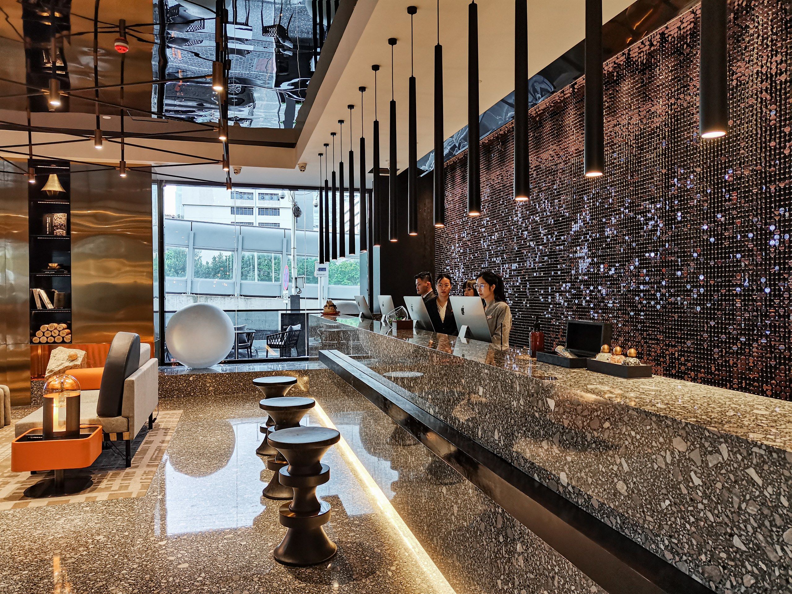 Een moderne hotellobby met een receptiebalie, een zithoek en een wand met decoratieve flessen.