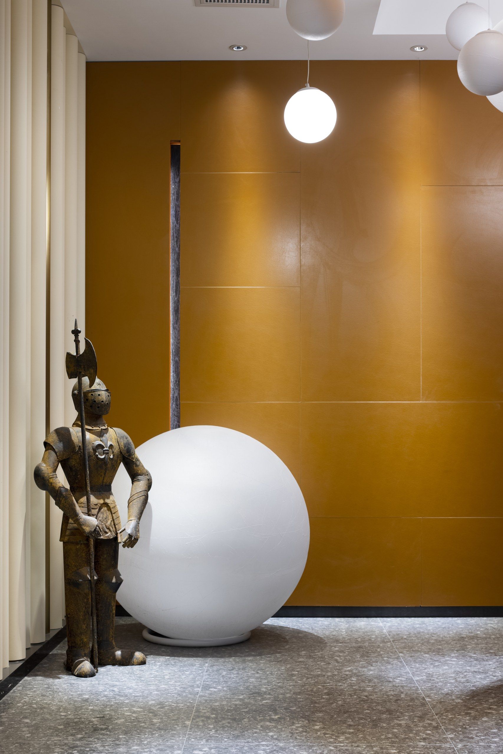 A statue of a knight holding a sword, a large white sphere, and a modern light fixture in a hallway with a yellow wall and white ceiling.