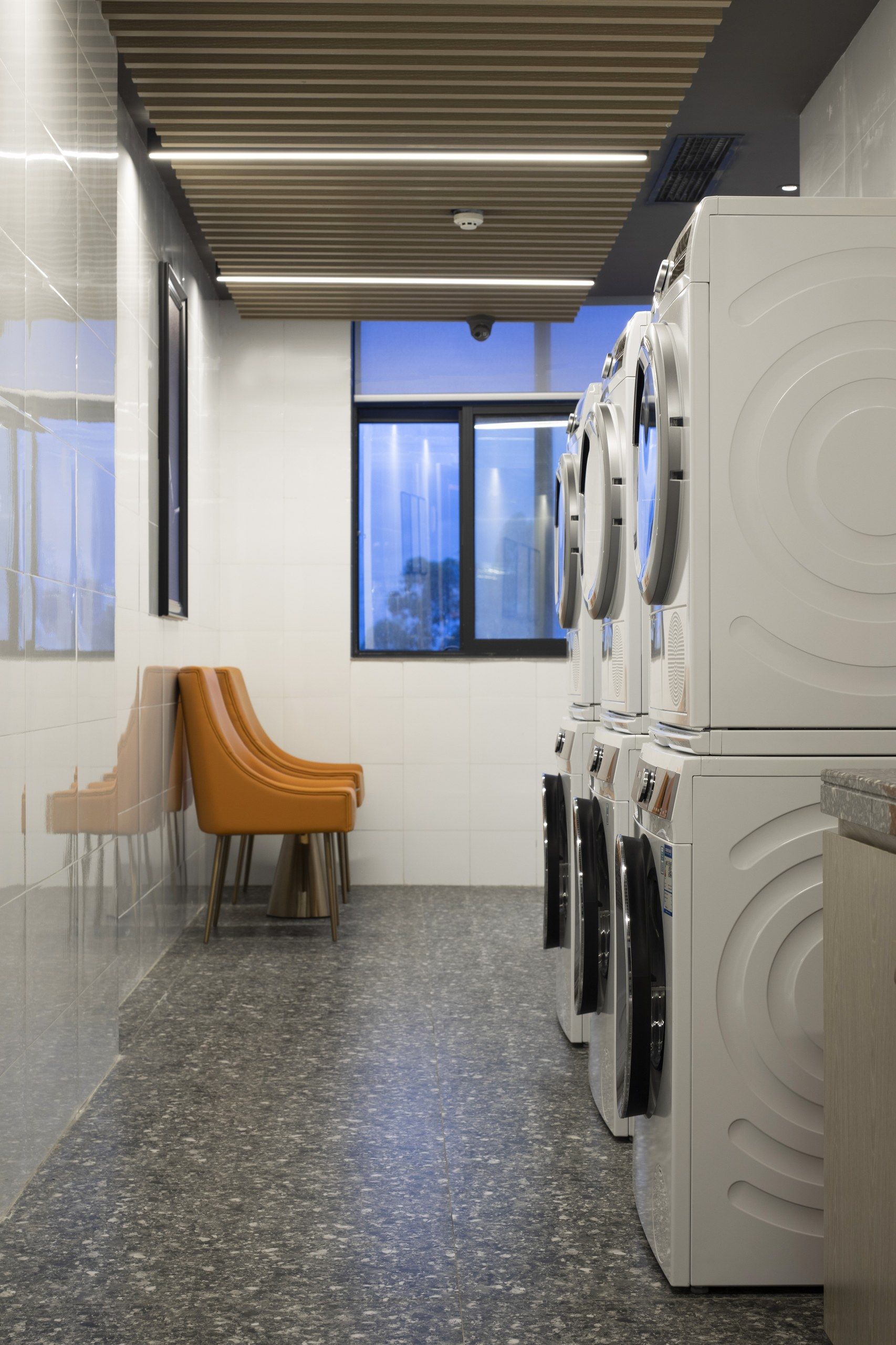 A laundry room with multiple washing machines, a seating area with chairs, and a large window providing a view.