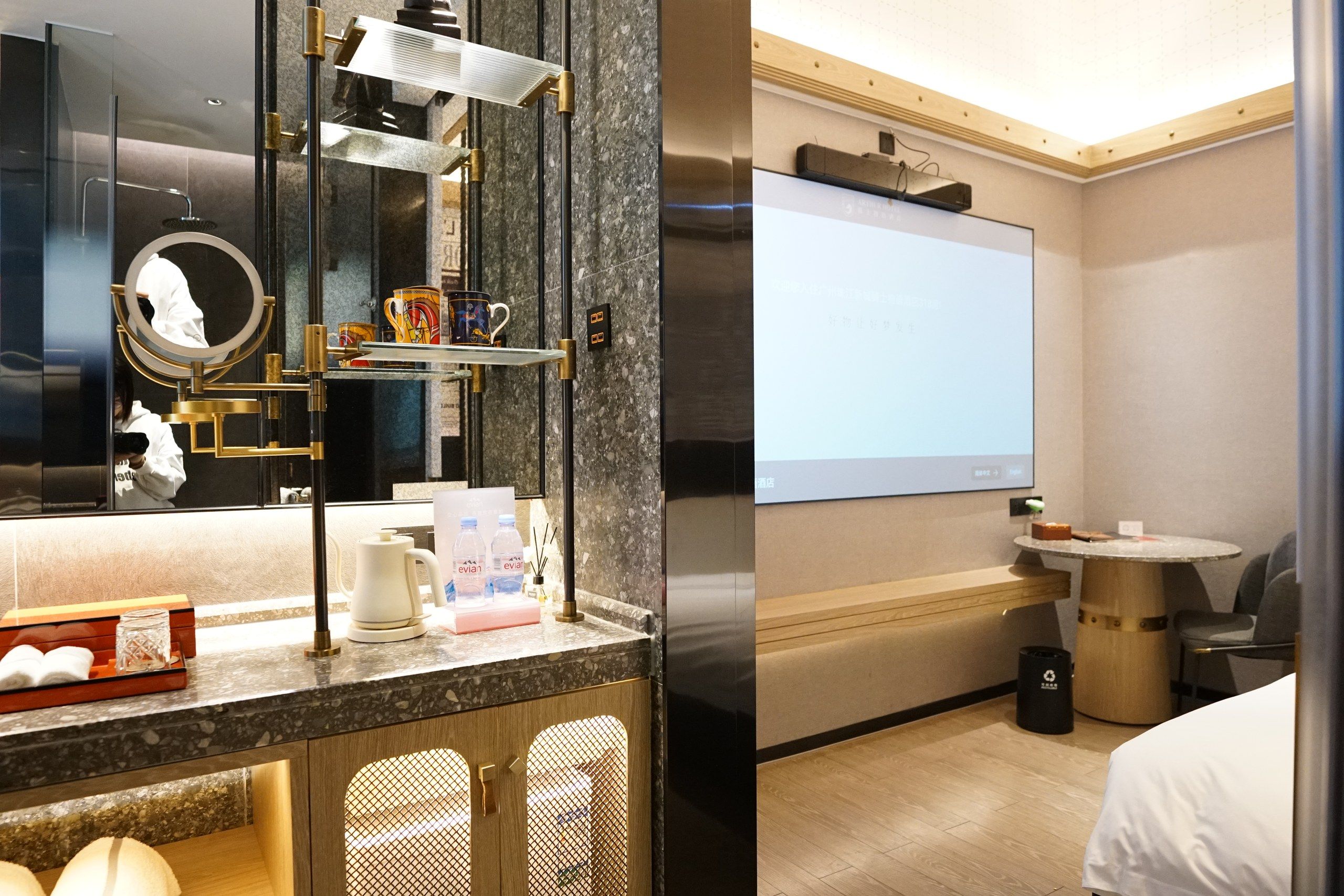 A hotel room with a large mirror, a coffee machine, a TV, and a small table with a chair.
