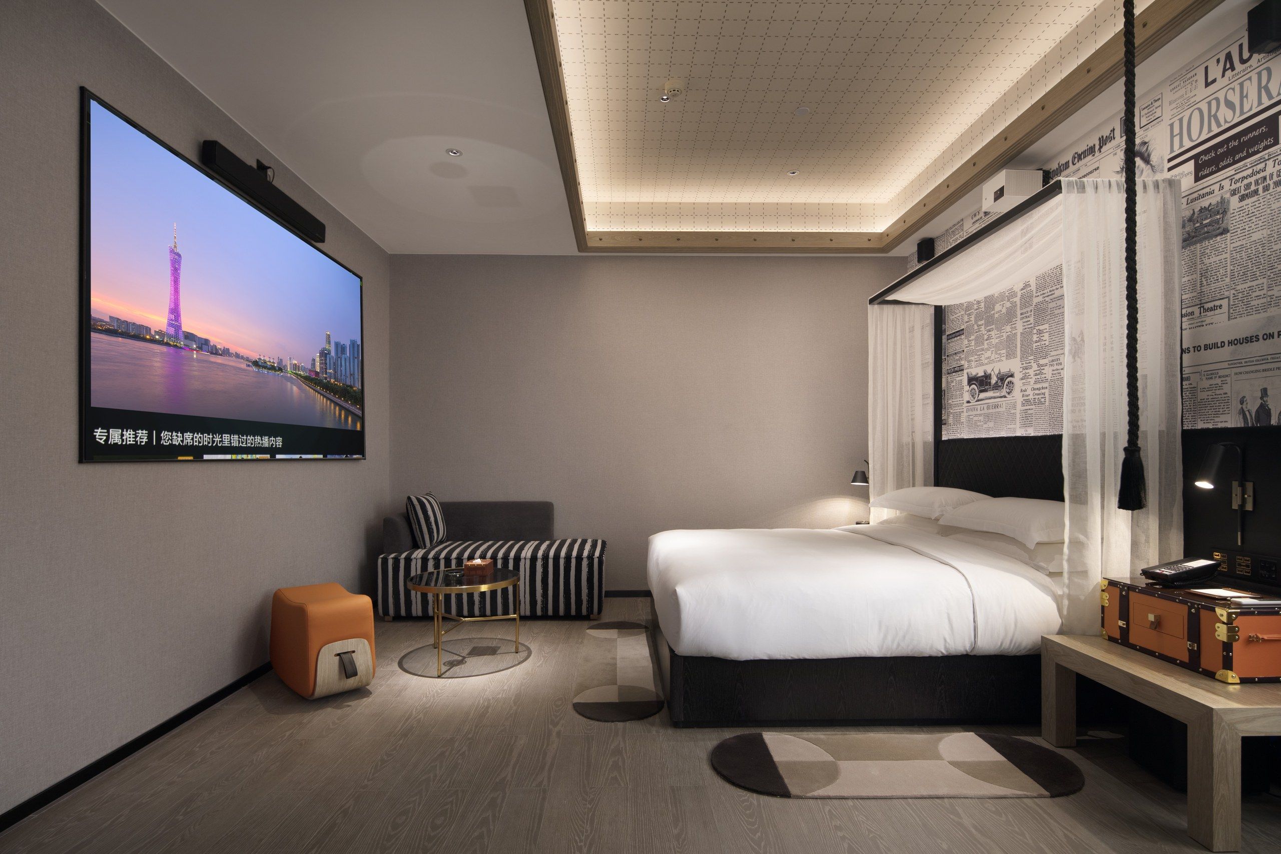 A modern hotel room with a large flat-screen TV, a bed, a sofa, a desk with a lamp, and a wall-mounted newspaper display.