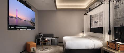 A modern hotel room with a large flat-screen TV, a bed, a sofa, a desk with a lamp, and a wall-mounted newspaper display.