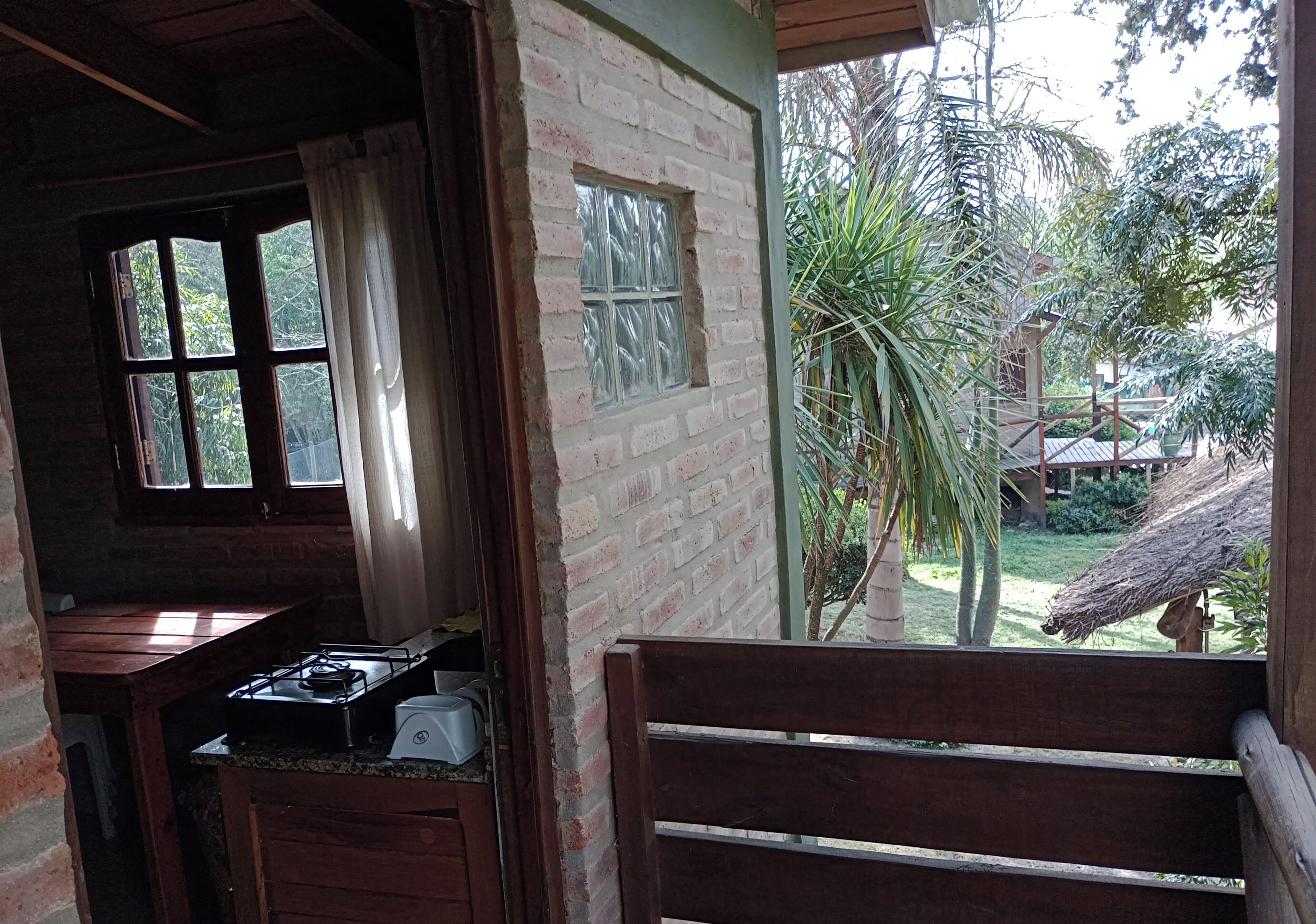 A rustic kitchen with a gas stove and a small sink, a wooden bench, and a view of a garden with palm trees and a thatched roof structure.