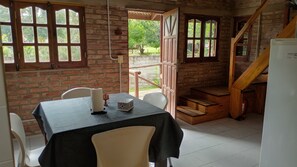 A room with a brick wall, a wooden staircase, a table with chairs, and a door leading outside.