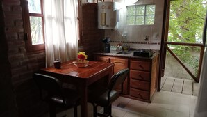 A kitchen with a brick wall, a window with curtains, a sink, a stove, and a dining area with a table and chairs. - Cabañas Refugio LAS ACEQUIAS (Tanti)