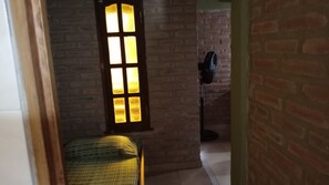 Una habitación con pared de ladrillo, ventana con marco de madera, un ventilador de pie y una cama con colcha verde a cuadros.