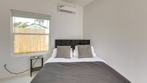 A bedroom with a bed, a window with blinds, an air conditioning unit, and a small bedside table. - #8 | South Padre Studio | Pool | Walk 2 Beach (South Padre Island)