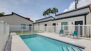 A swimming pool with a white fence, two chairs, and a building with a garage door in the background. - #8 | South Padre Studio | Pool | Walk 2 Beach (South Padre Island)