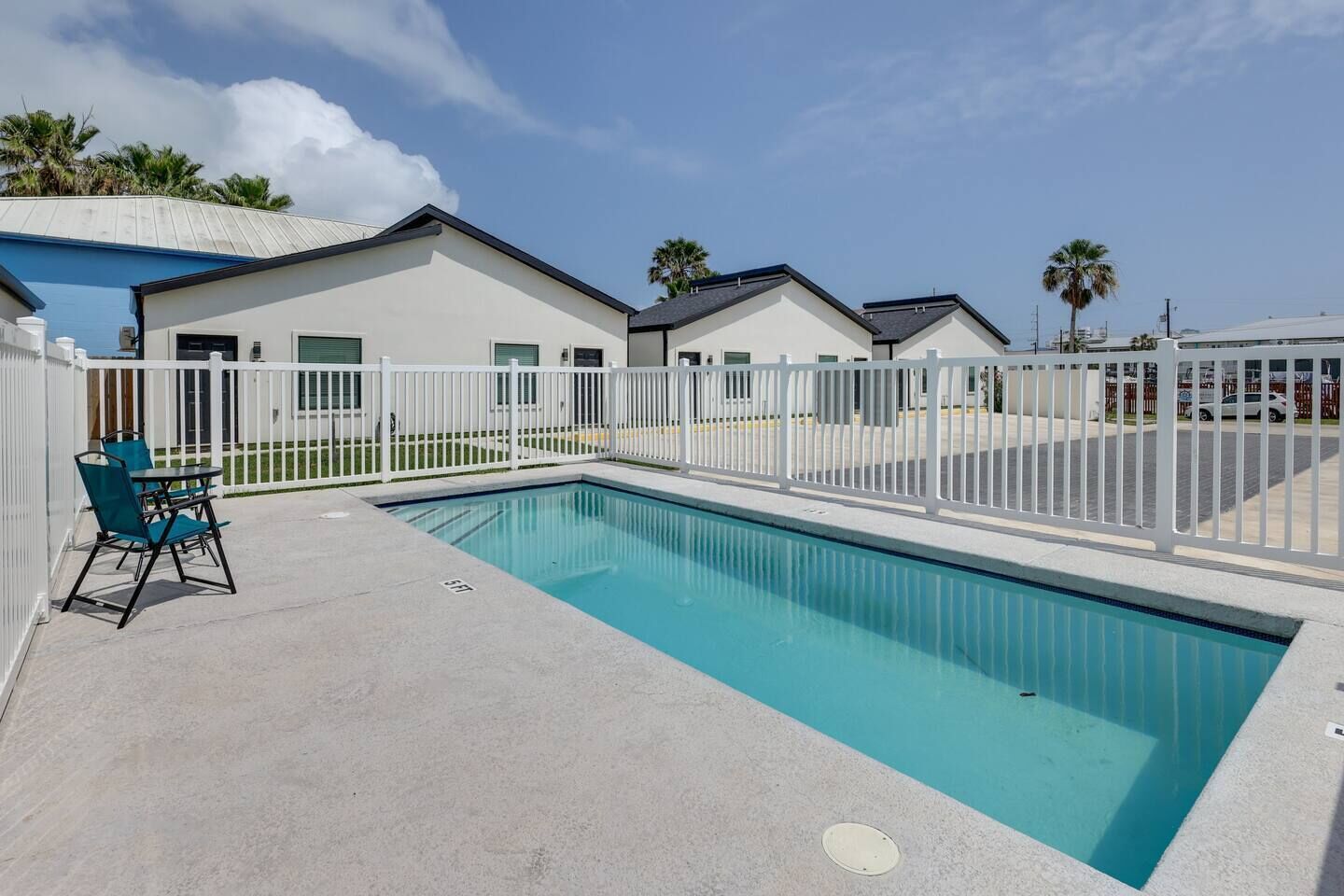 Une piscine entourée d’une clôture blanche, une table avec des chaises, et un bâtiment avec un toit plat en arrière-plan.
