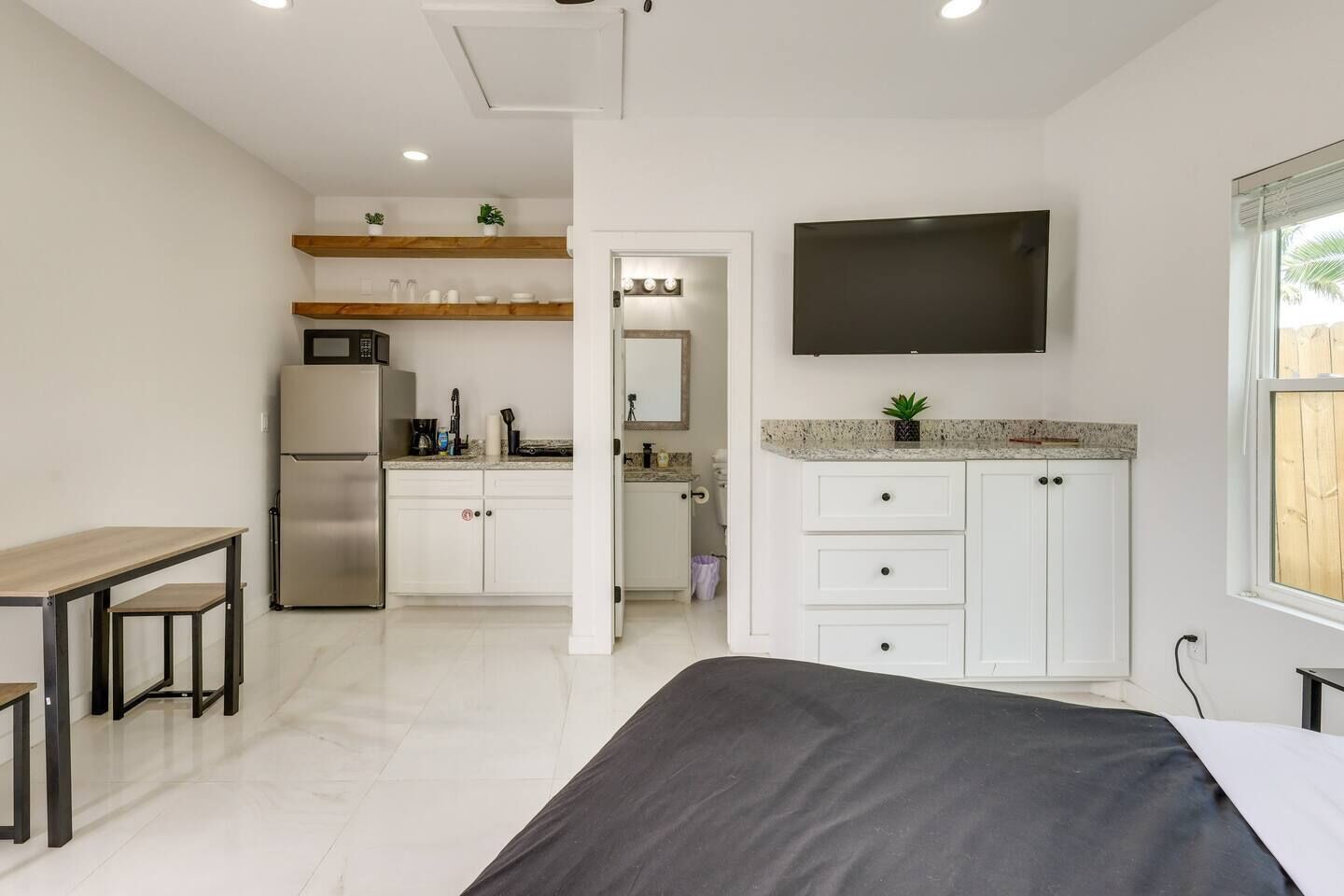 A compact living space with a kitchenette, a flat-screen TV mounted above the counter, a bed with a dark cover, and a small table with chairs.