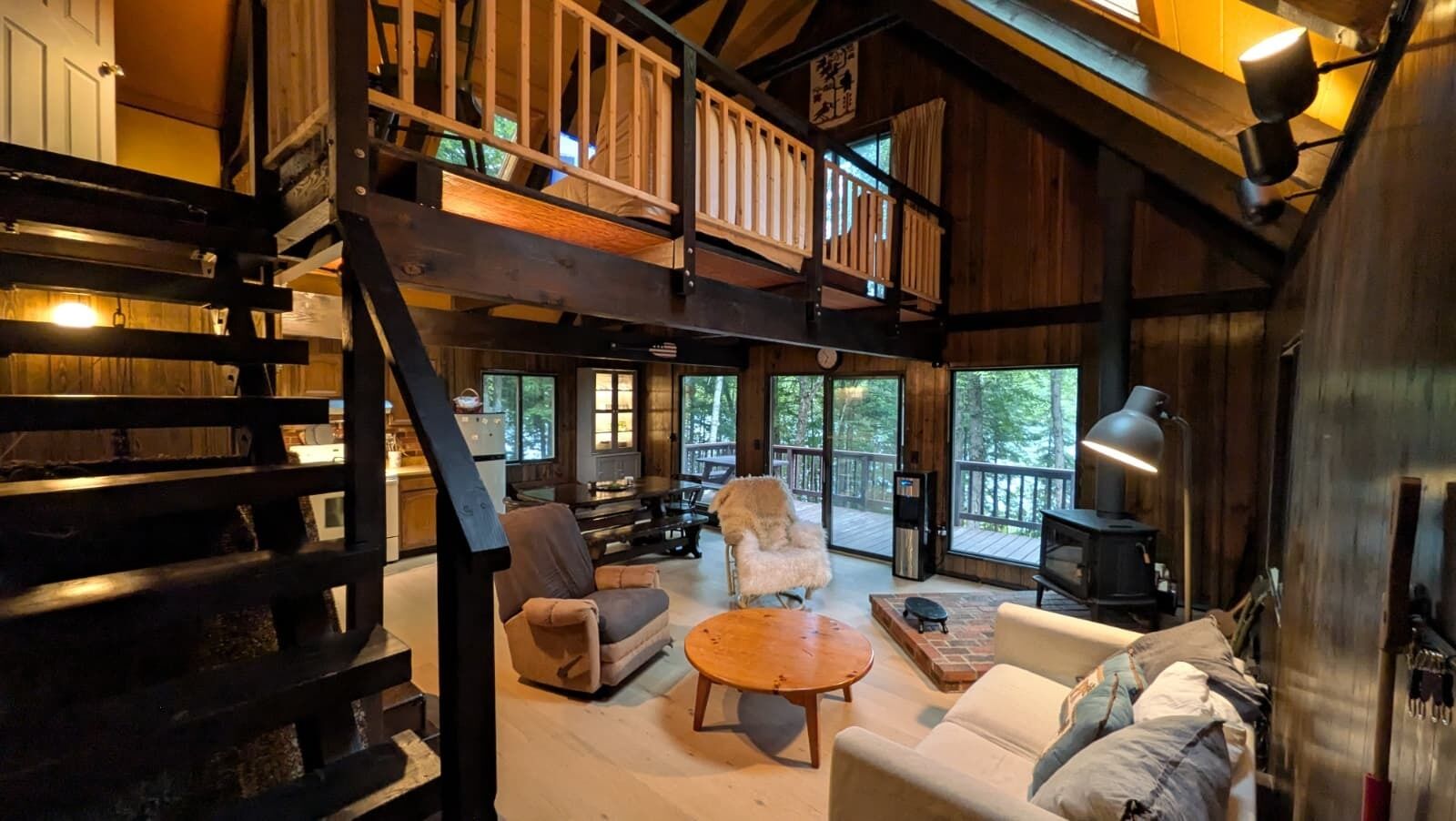 A cozy cabin interior with a wooden staircase, a fireplace, and a balcony overlooking a forest.