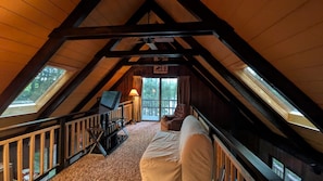 A cozy attic bedroom with a sloped ceiling, a bed, a TV, and a window overlooking a deck.