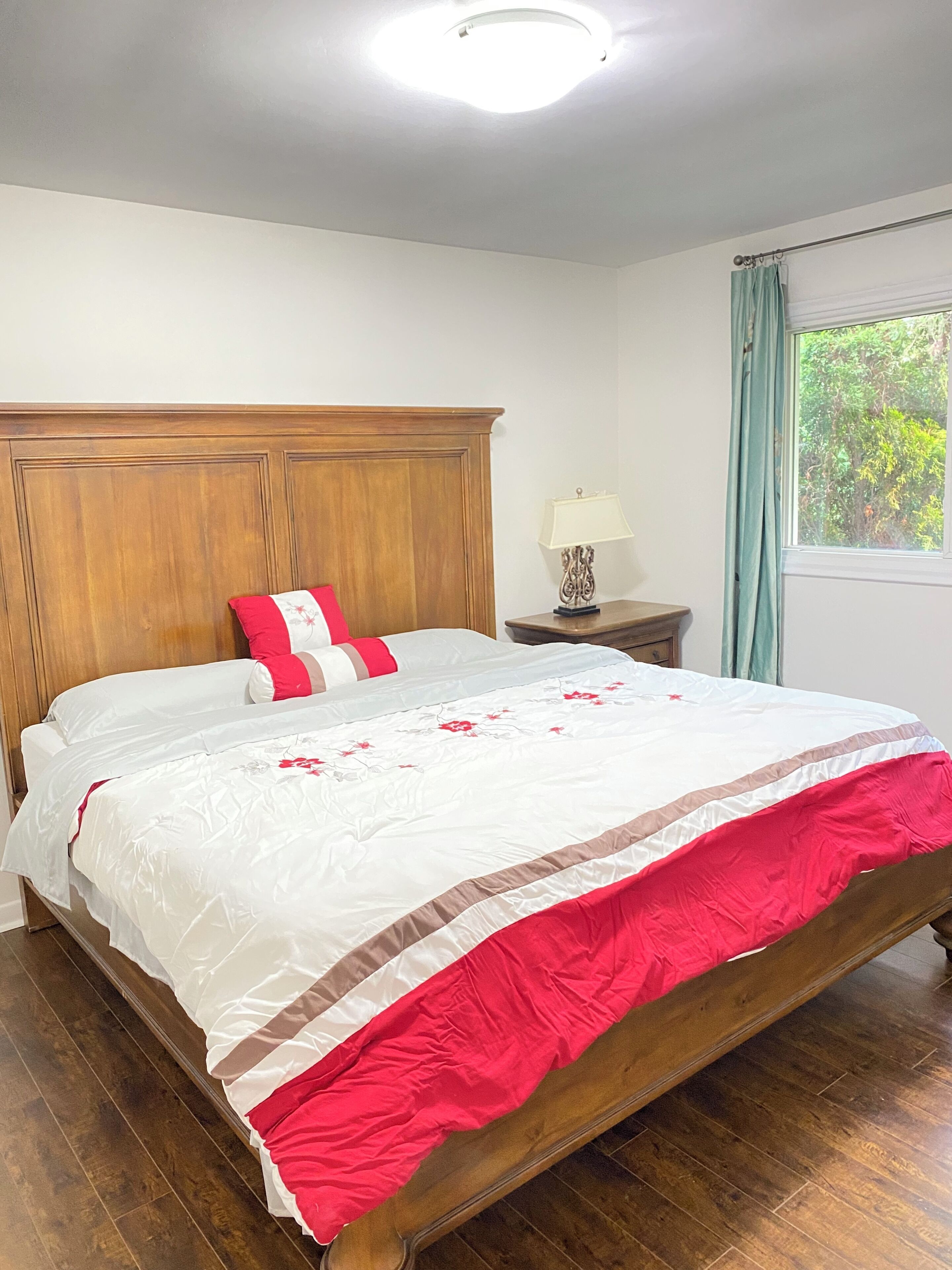 A bedroom with a wooden headboard, a bed with a white and red bedspread, a nightstand with a lamp, and a window with curtains.