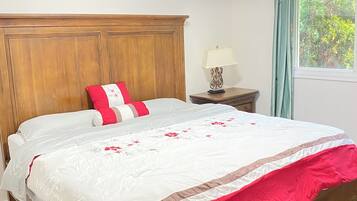 A bedroom with a wooden headboard, a bed with a white and red bedspread, a nightstand with a lamp, and a window with curtains.