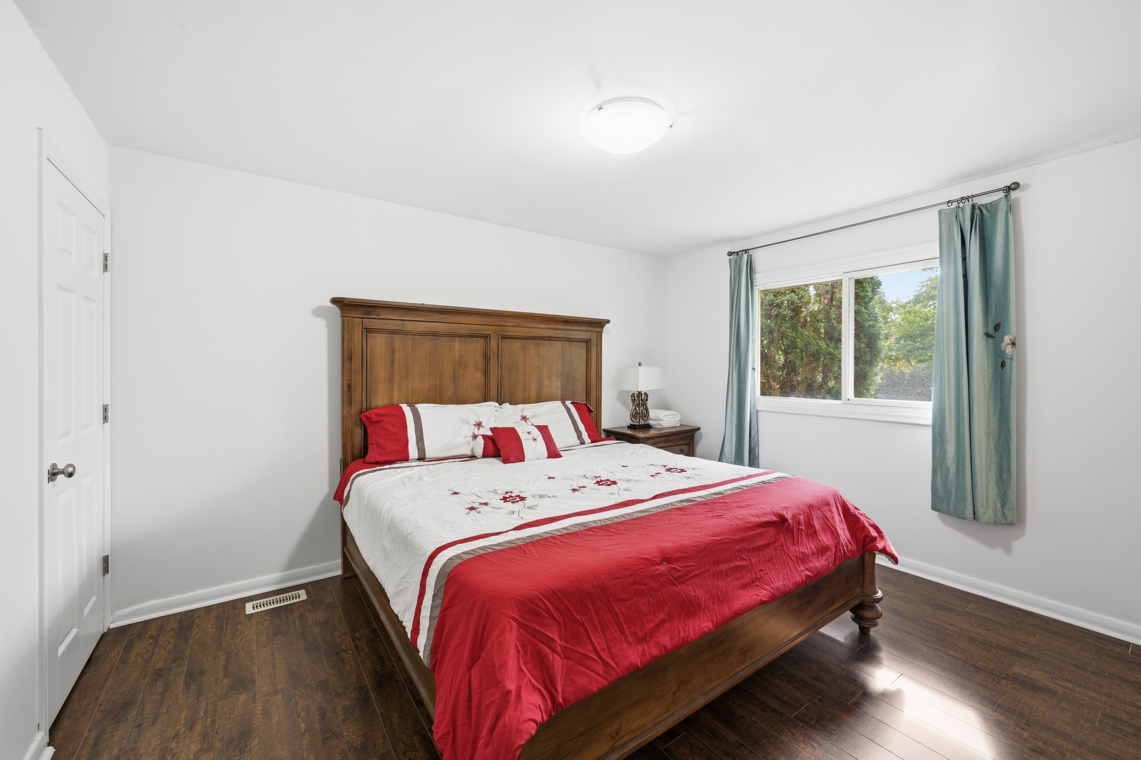 A bedroom with a wooden bed, white walls, a window with curtains, and a door.