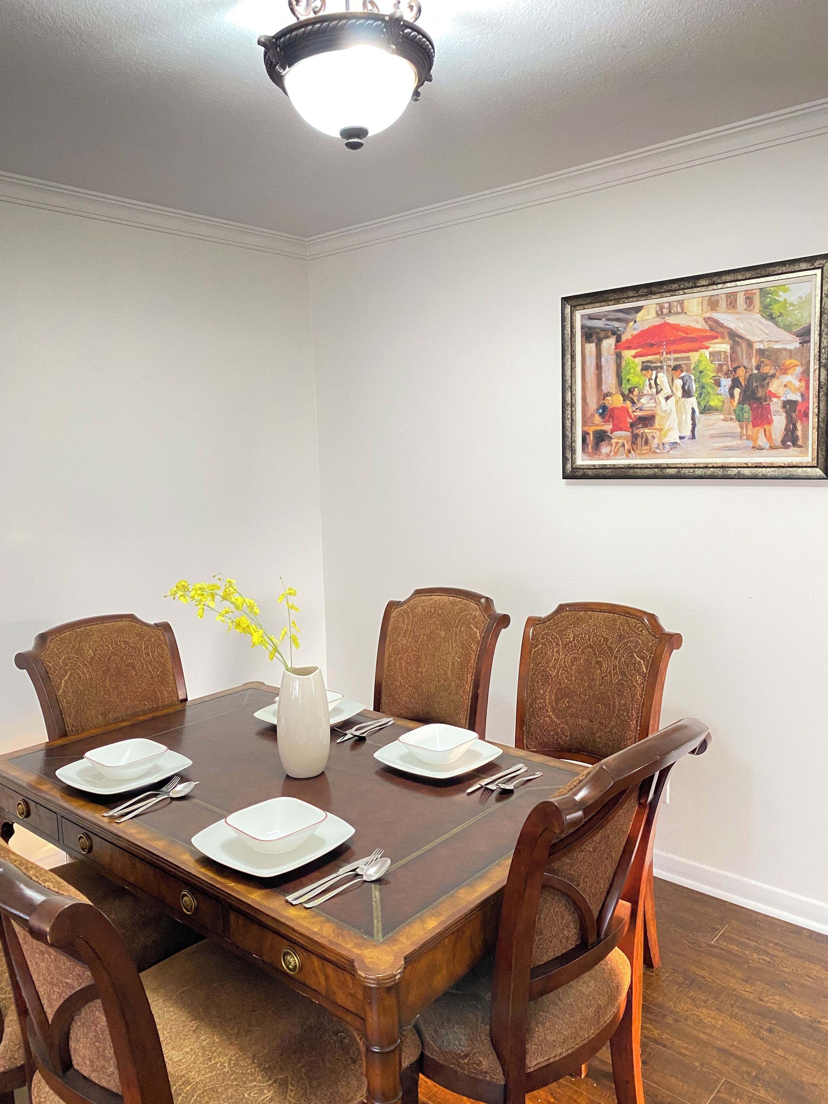 A dining room with a rectangular table set for four, surrounded by upholstered chairs, a vase with yellow flowers, and a framed painting on the wall.