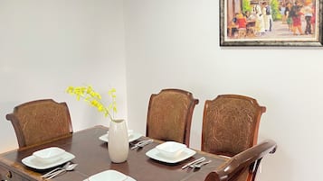 A dining room with a rectangular table set for four, surrounded by upholstered chairs, a vase with yellow flowers, and a framed painting on the wall.