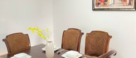 A dining room with a rectangular table set for four, surrounded by upholstered chairs, a vase with yellow flowers, and a framed painting on the wall.