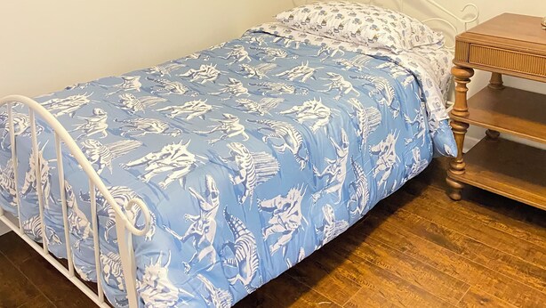 Una cama individual con estructura metálica y colcha azul estampada en una habitación con piso de madera y pared lisa.