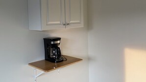Una habitación con un armario blanco sobre una estantería de madera, una cafetera negra en la estantería y un suelo de tono claro.