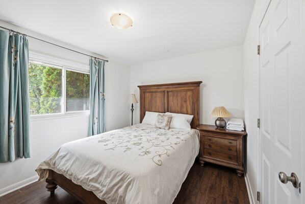 A bedroom with a wooden bed, a nightstand with a lamp, and a window with curtains. - cozy three bedroom detached house with free parking (Toronto)