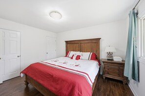 Un dormitori amb un llit de fusta, parets blanques, una nitrera de fusta i una finestra amb cortines.