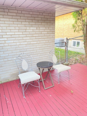 Un espacio de estar al aire libre con techo, una pared de ladrillo, una mesa de metal y dos sillas blancas con cojines.