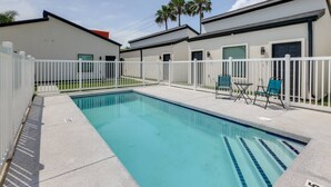 A swimming pool with a white fence, two chairs, and a building with a garage door. - #9 | Beach Escape Studio w/Pool | Walk Everywhere (South Padre Island)