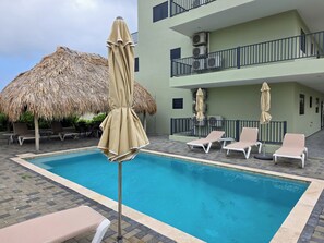 A swimming pool with lounge chairs and umbrellas, a thatched roof structure, and a multi-story building with balconies and air conditioning units. - Elisabeth Beach House near Mambo Beach (Willemstad)