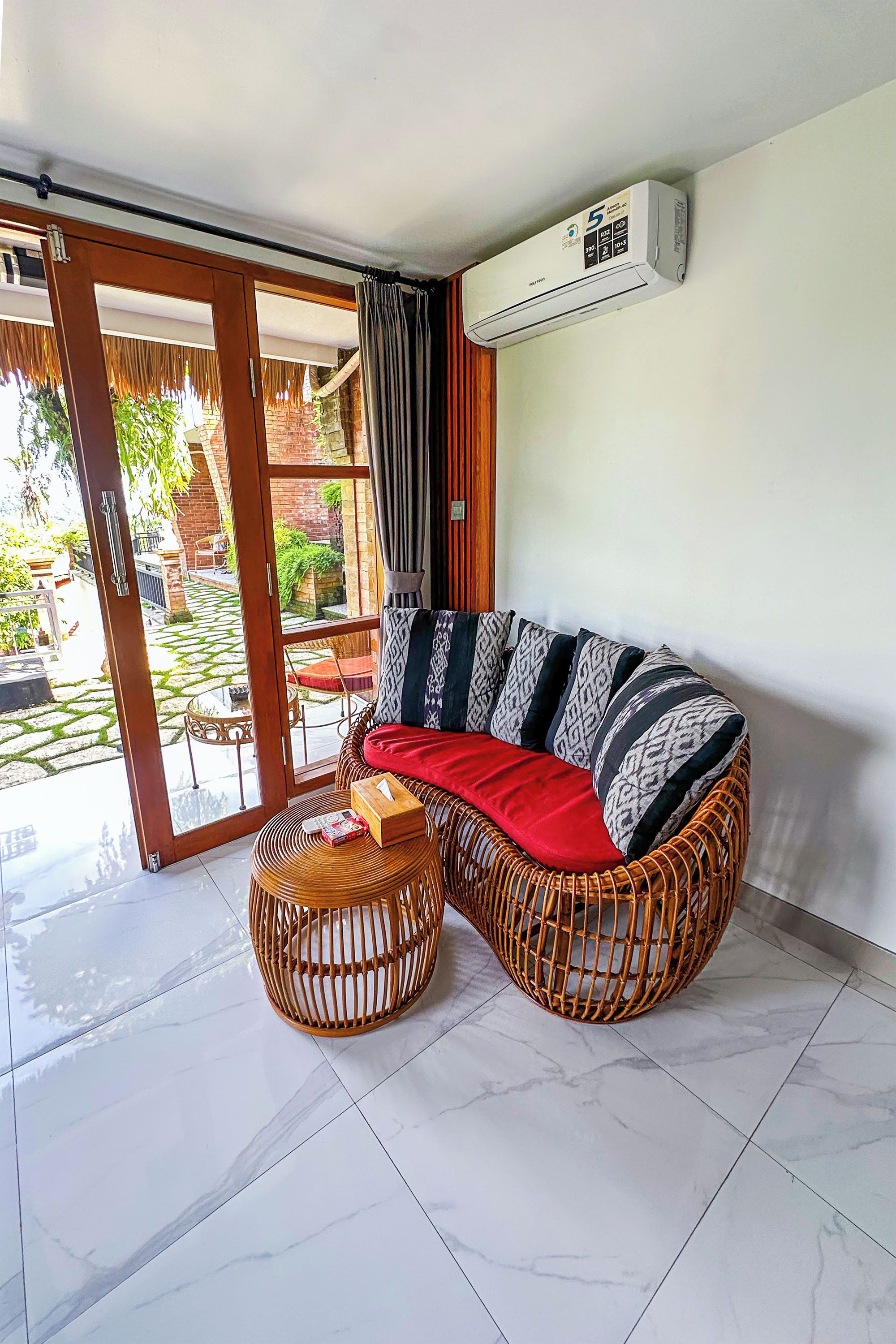 Una sala moderna con un sofá seccional, dos sillones de mimbre, un ventanal que da a un patio y un equipo de aire acondicionado en la pared.
