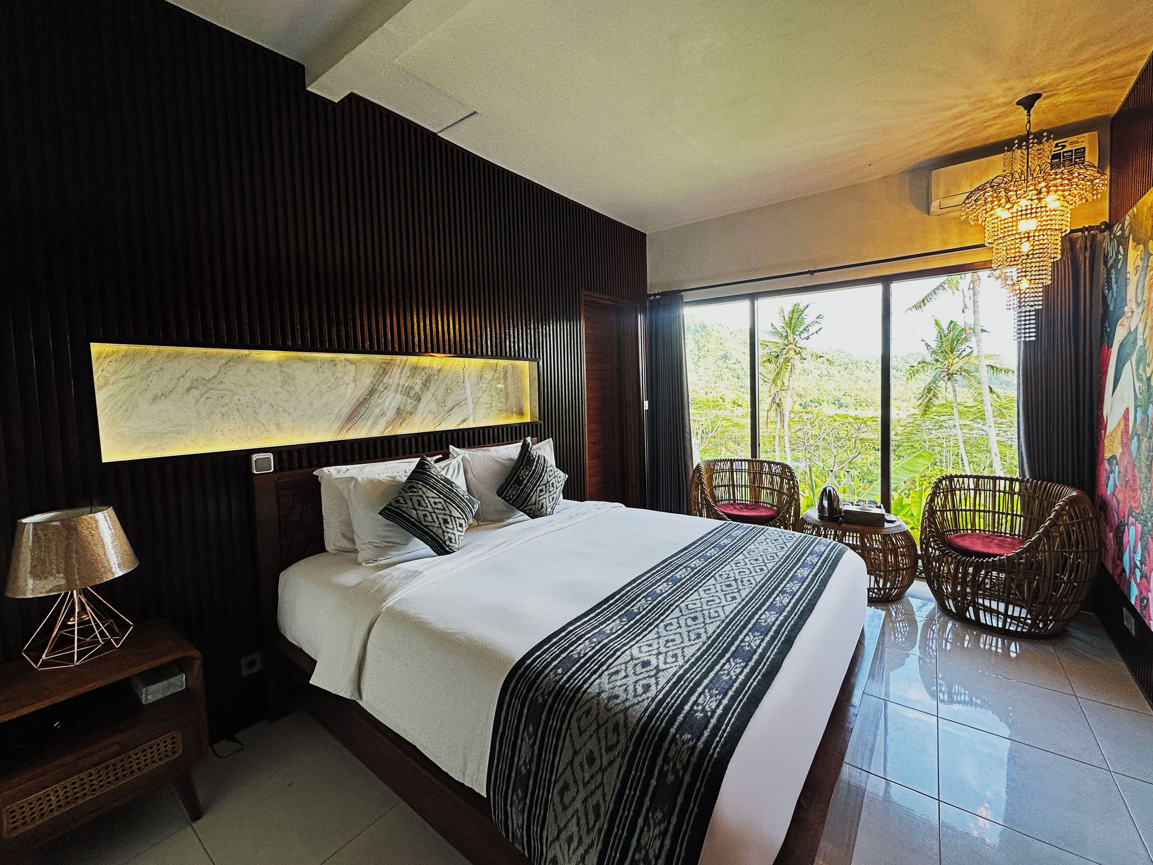 Habitación de hotel con una cama grande, cabecera con diseño tipo mármol, vista a palmeras a través de ventanas amplias y un candelabro.