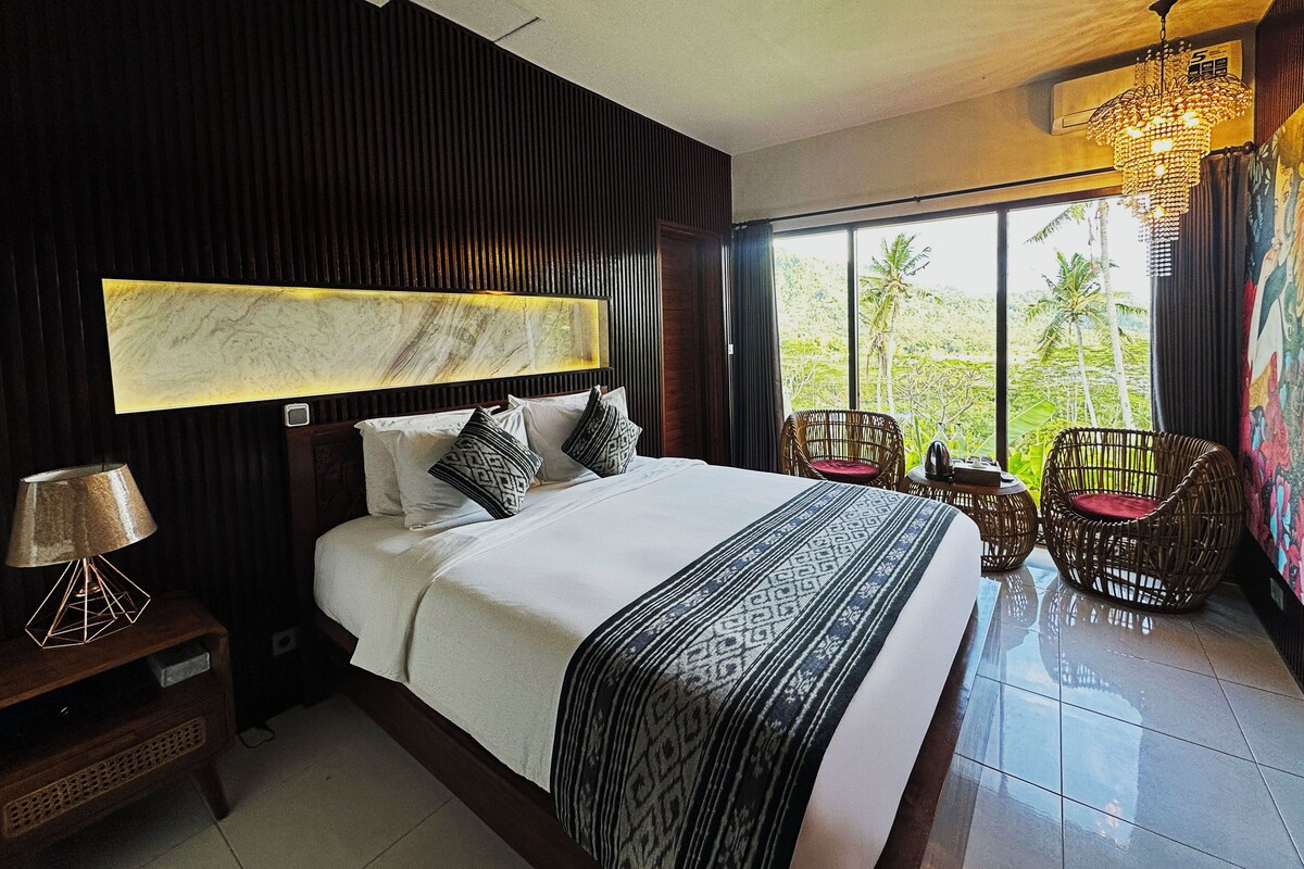 A hotel room with a large bed, a headboard with a marble-like design, a view of palm trees through large windows, and a chandelier.
