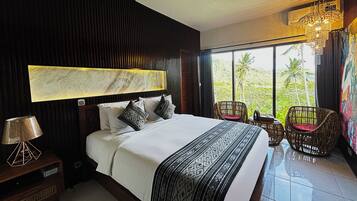 Habitación de hotel con una cama grande, cabecera con diseño tipo mármol, vista a palmeras a través de ventanas amplias y un candelabro.