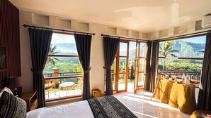 A hotel room with a large bed, a balcony with wicker furniture, and a view of a lush landscape. - Aura Cemapaka (Karangasem)