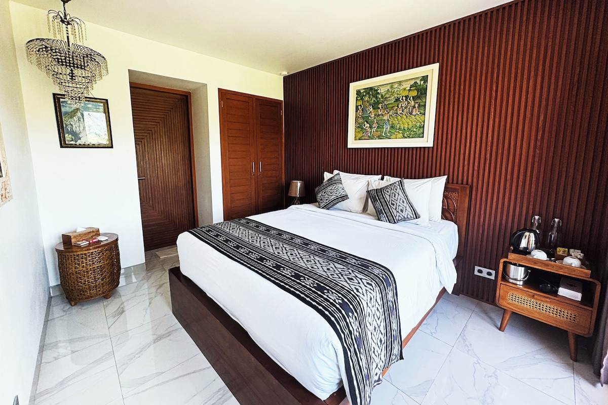 A bedroom with a large bed, a wooden wardrobe, a wicker side table, and a framed picture on the wall.