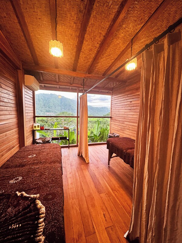 A wooden room with a bed, a massage table, and a view of a lush landscape.