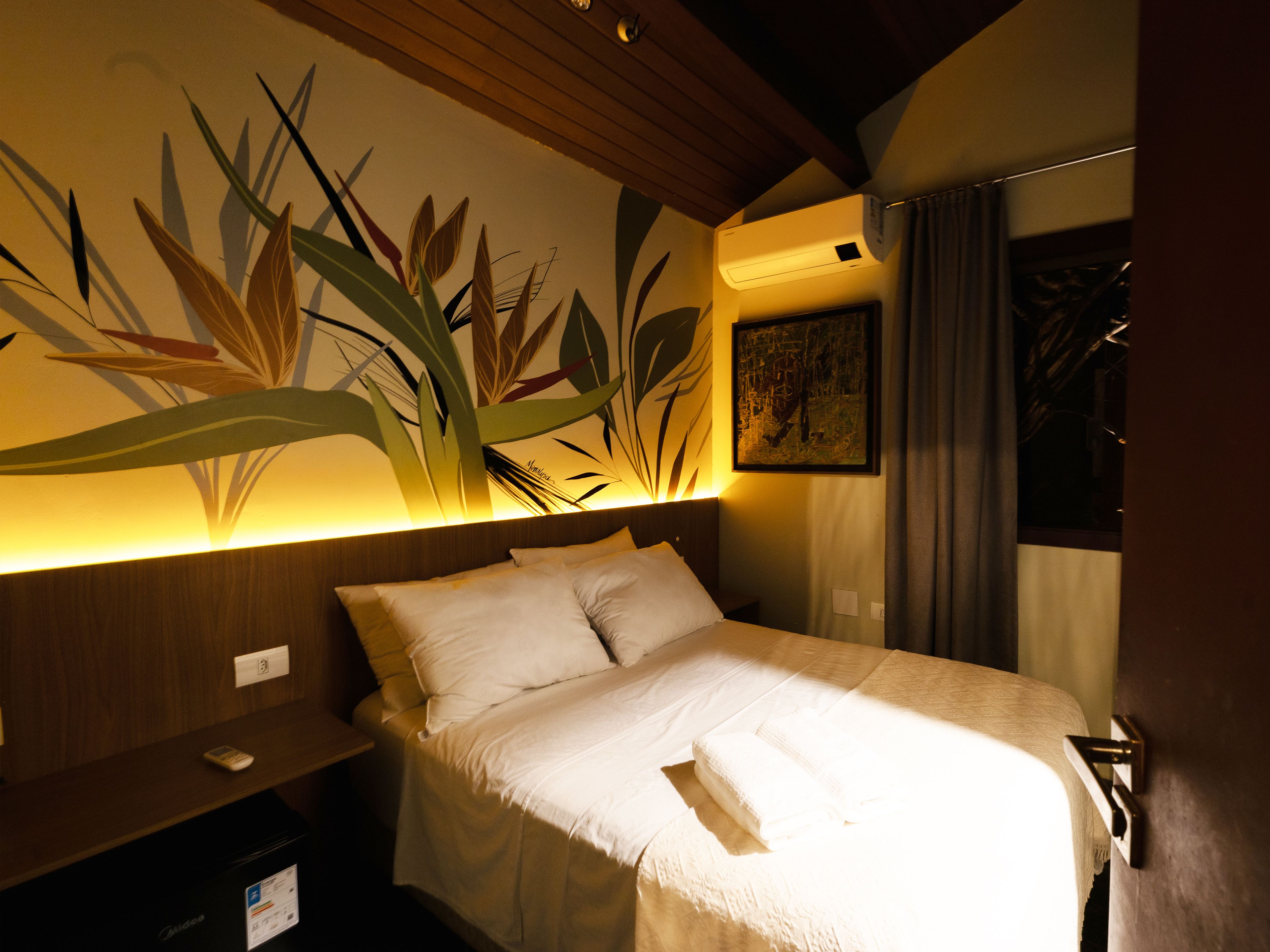 Quarto de hotel com cama, mesa de cabeceira, ar-condicionado fixado na parede e uma pintura.