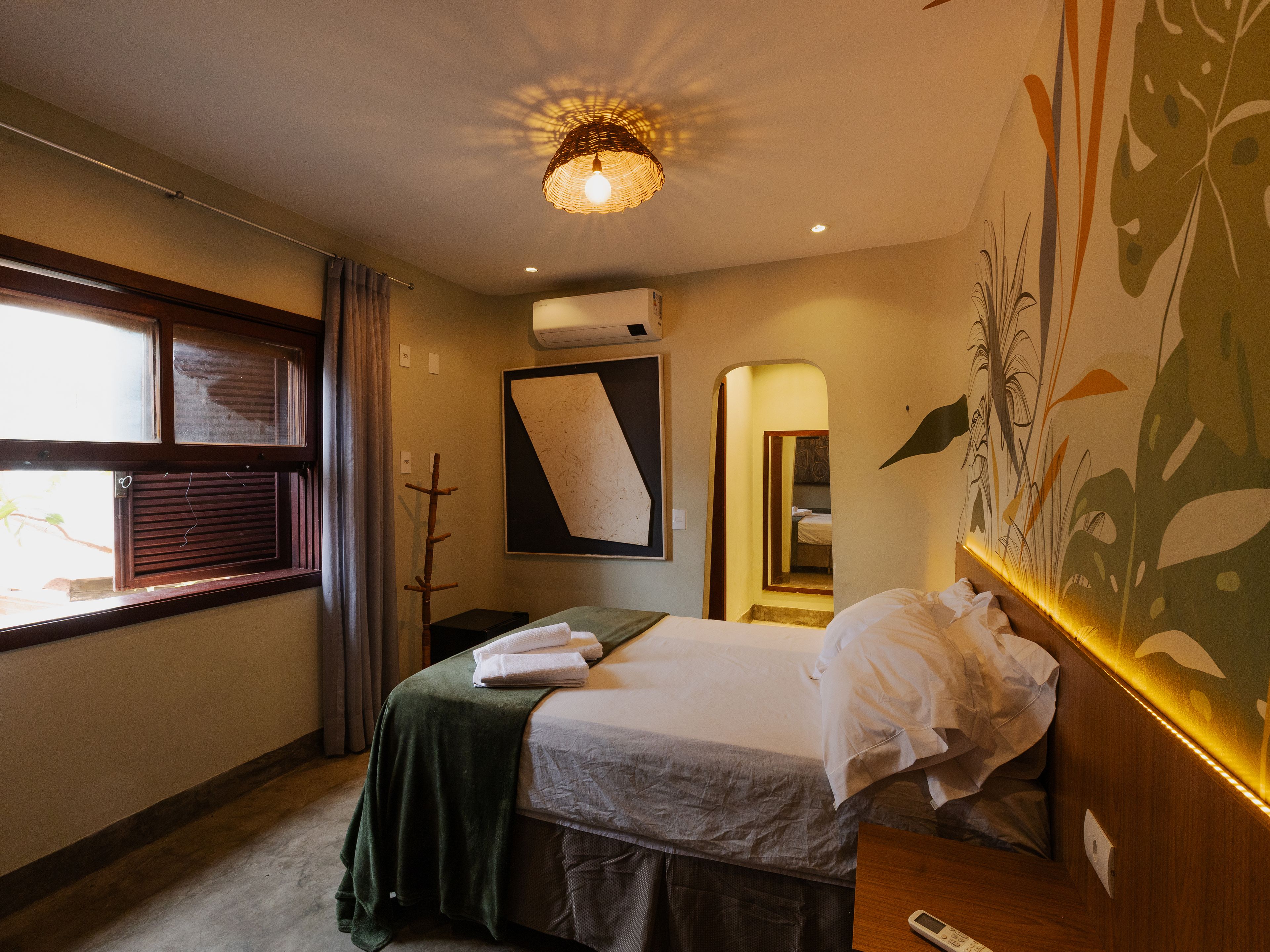 A hotel room with a bed, a window with curtains, a wall-mounted air conditioner, and a ceiling light fixture.