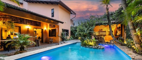 Una piscina con área de patio, rodeada de abundante vegetación y un edificio con asientos al aire libre.