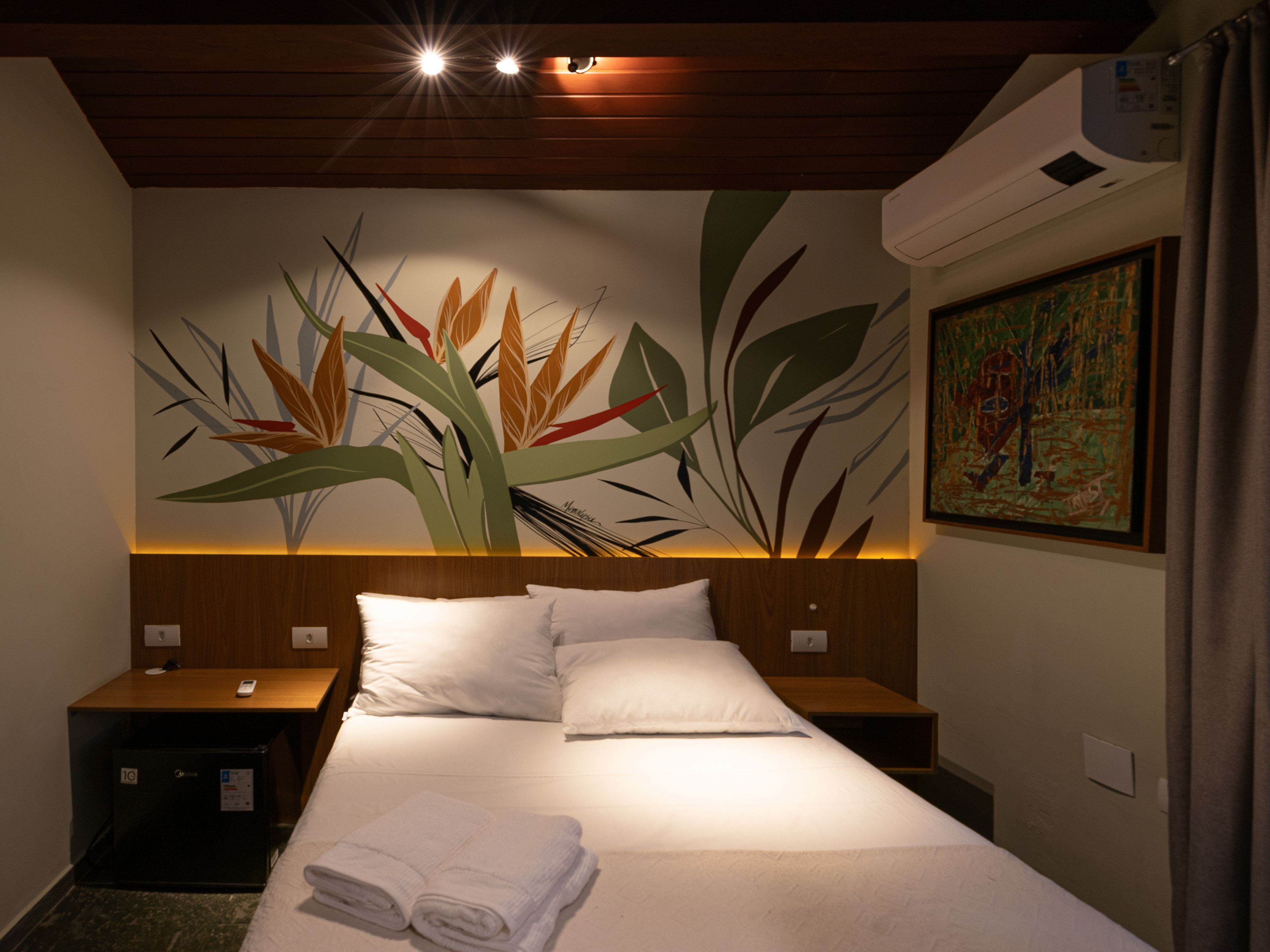 A hotel room with a bed, two bedside tables, a wall-mounted air conditioner, and a painting.