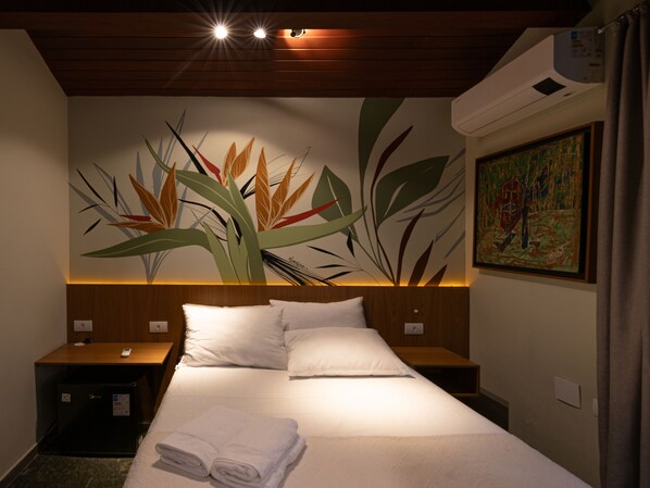A hotel room with a bed, two bedside tables, a wall-mounted air conditioner, and a painting.
