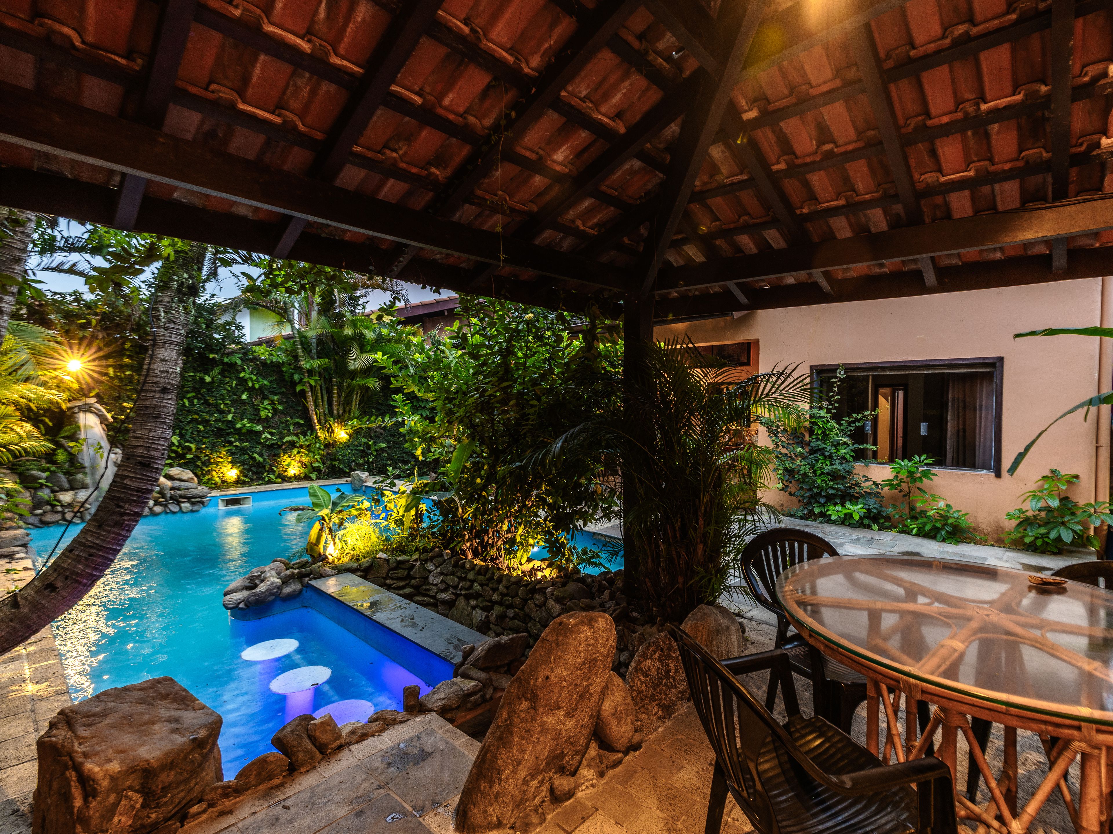 Uma área de piscina com mesa e cadeiras de vidro, cercada por pedras e plantas.