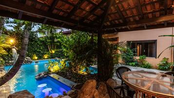 Uma área de piscina com mesa e cadeiras de vidro, cercada por pedras e plantas.